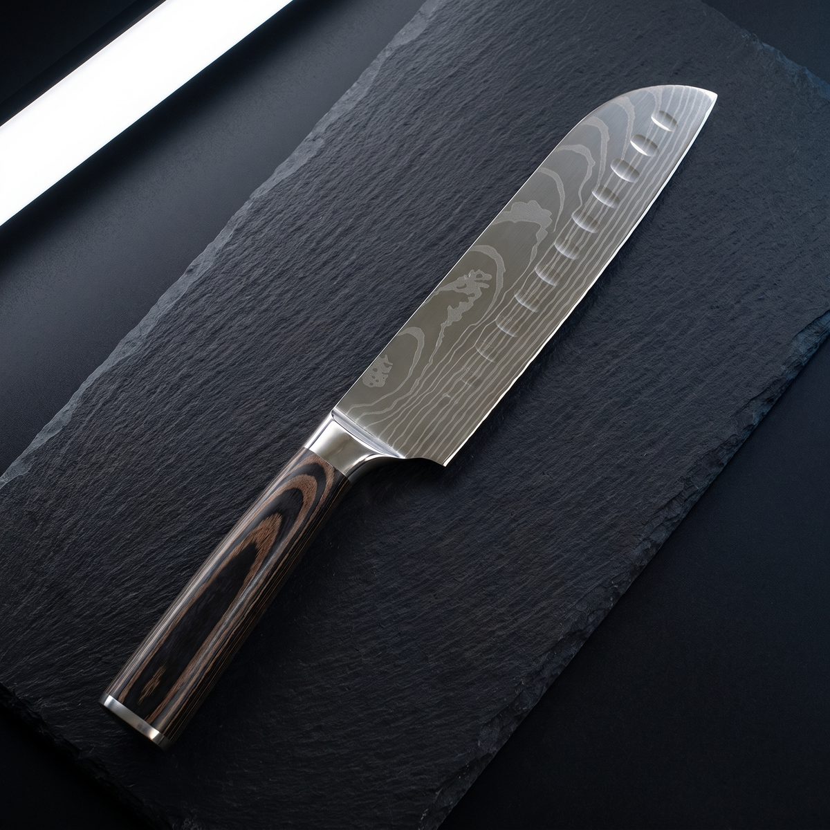 Couteau santoku TitaniumCook mit wellenförmig gemusterter Klinge und schwarz-braun gemasertem Griff, auf schwarzem Schiefer.